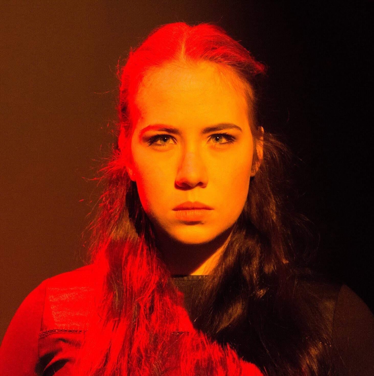 A heavily red-tinged head and shoulders photo of a young white woman with long brown hair. She is facing the camera with a challenging expression.